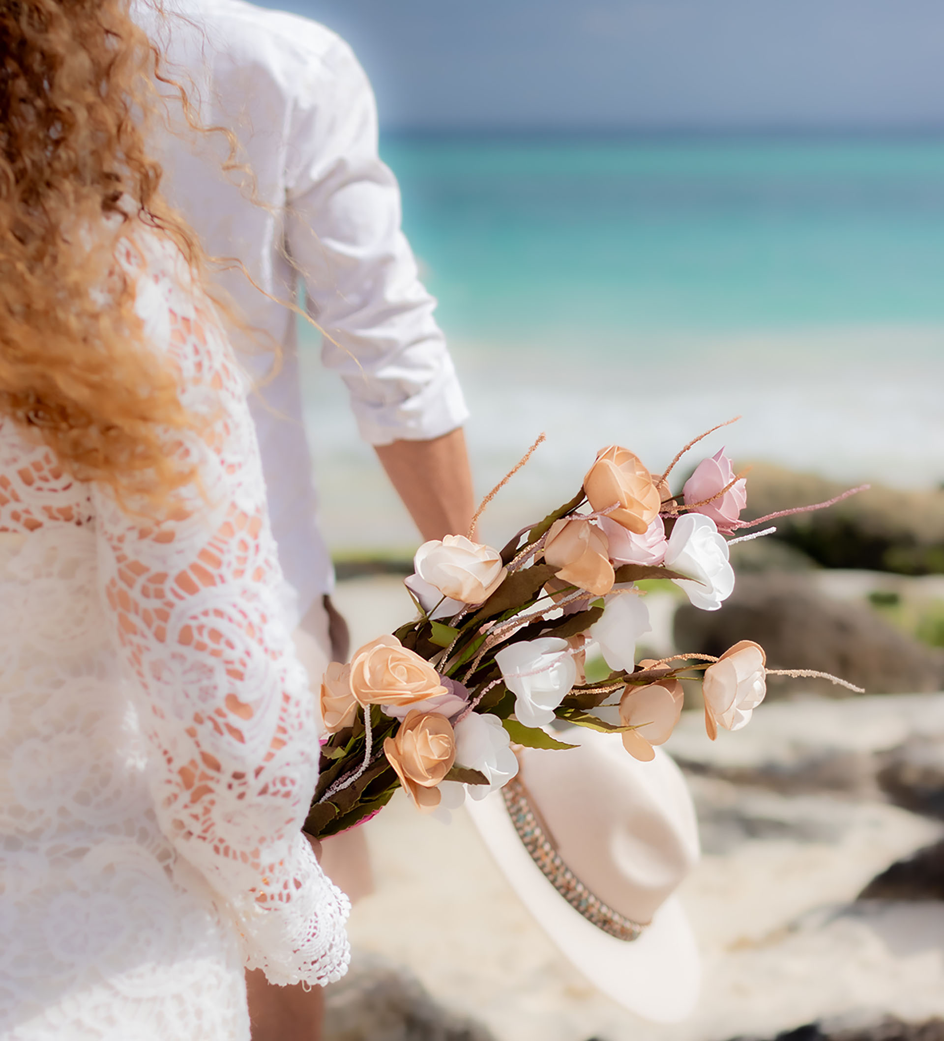 Primer plano de novia de espaldas con vestido de encaje blanco sosteniendo un ramo de flores en tonos pastel frente al mar turquesa de Playa del Carmen.