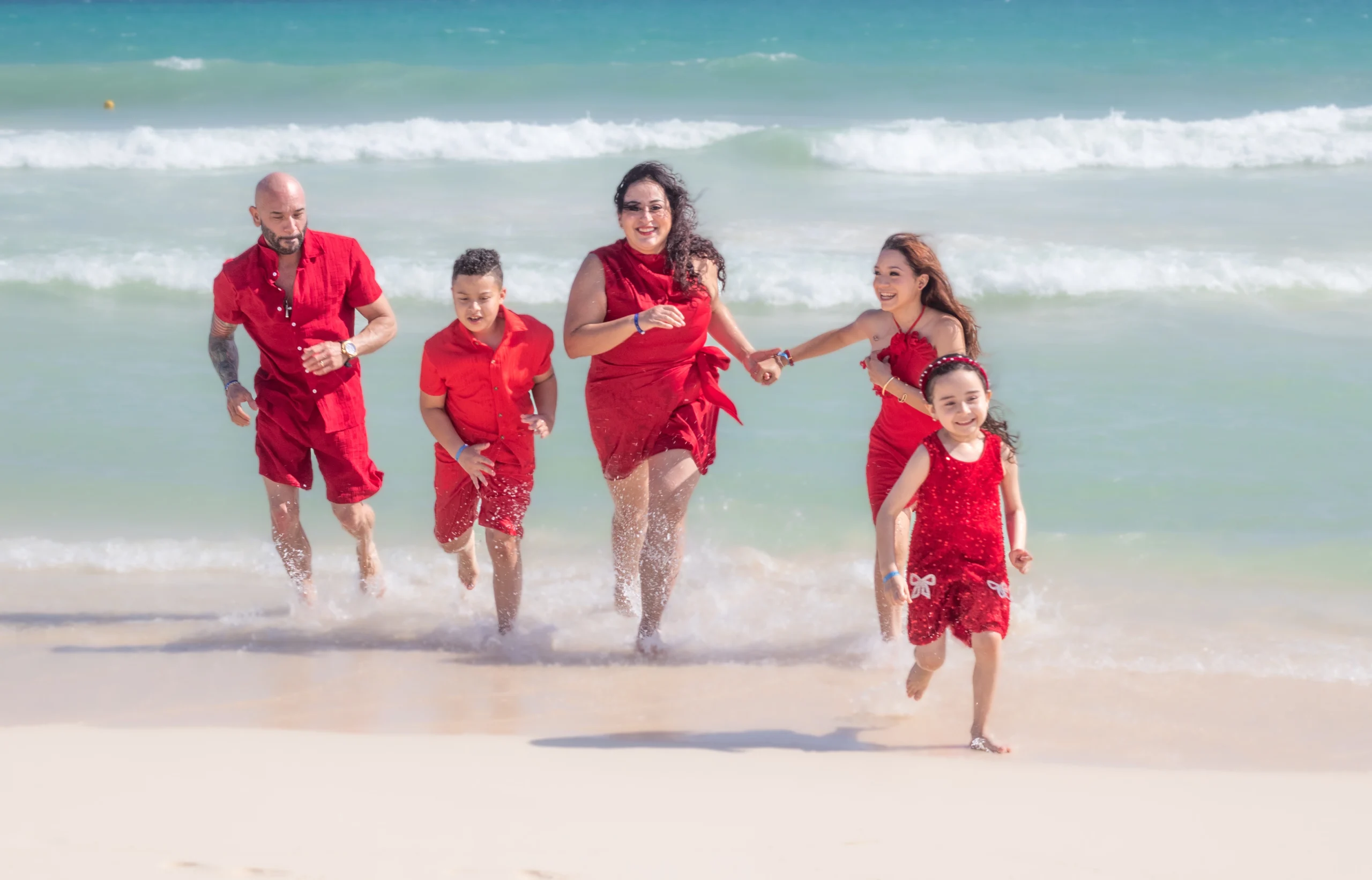 Familia feliz de cuatro integrantes sonriendo y abrazados en la orilla de la playa con el mar azul de fondo.