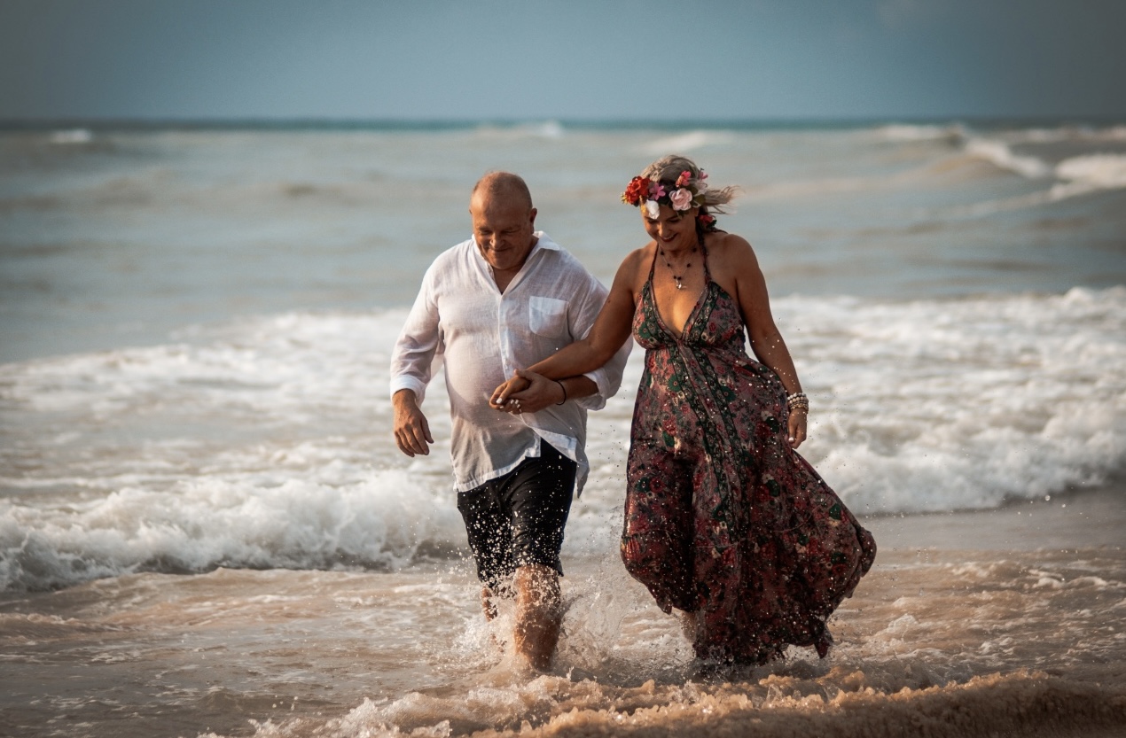 Pareja madura caminando de la mano por la orilla del mar en Playa del Carmen; ella lleva un vestido floral y corona de flores, él camisa blanca, con olas de fondo.